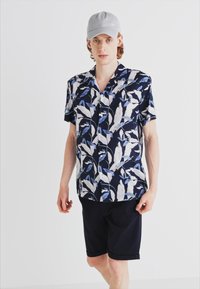 Camicia a maniche corte di colore blu navy con un motivo floreale blu e bianco; abbinata a pantaloni corti scuri; chi la indossa ha anche un berretto grigio chiaro.