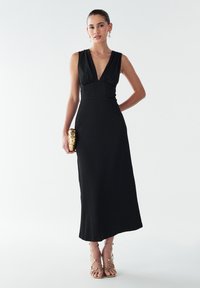 Robe maxi sans manches noire avec un décolleté en V profond et un tissu texturé. Le mannequin tient une pochette décorative dorée et porte des talons dorés à lanières.