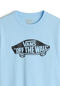 Světle modré tričko vyrobené z bavlny s kulatým výstřihem. Obsahuje černou grafiku skateboardu a text "Vans 'Off The Wall'."