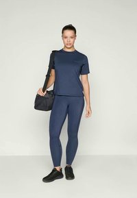 Tricou sportiv de culoare bleumarin și colanți din material elastic, asortați cu o geantă neagră de umăr. Model purtând adidași negri.