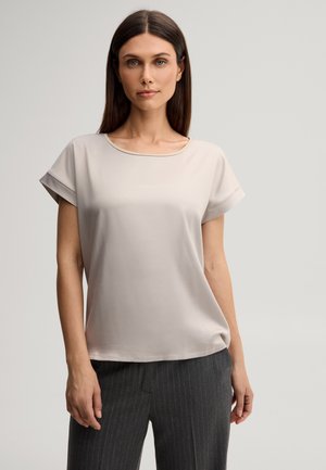 Vrouw met recht bruin haar, gekleed in een lichtbeige blouse met korte mouwen en donkergrijze pantalon met fijne strepen tegen een effen achtergrond.