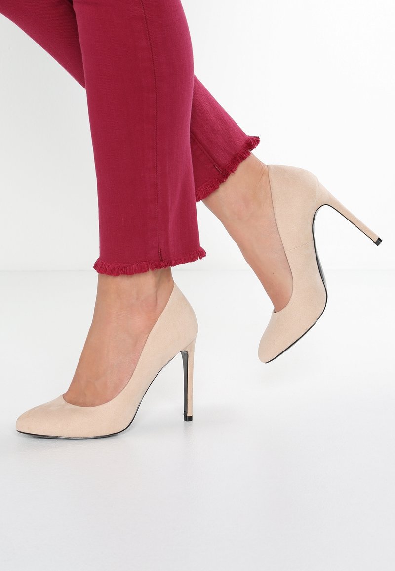 Escarpins beige en daim avec une texture lisse, un bout pointu et un talon noir élégant, associés à un pantalon bordeaux effiloché coupé au-dessus de la cheville.