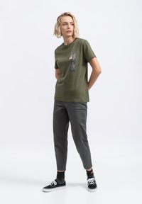 Olijfgroene t-shirt met een grafiek van een boom en een dier op de borst, gecombineerd met grijze tapered broek en zwarte sneakers. Soepele textuur.