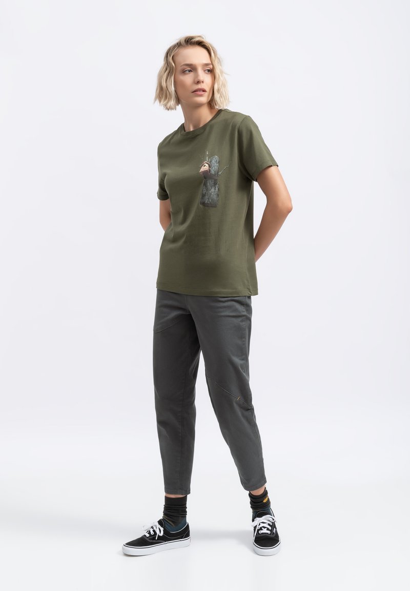 Olijfgroene t-shirt met een grafiek van een boom en een dier op de borst, gecombineerd met grijze tapered broek en zwarte sneakers. Soepele textuur.