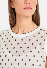 Weißes, durchsichtiges Oberteil mit runder Ausschnitt, verziert mit einer gewellten Borte und mehrfarbigen Punkten in Pink, Braun und Orange.