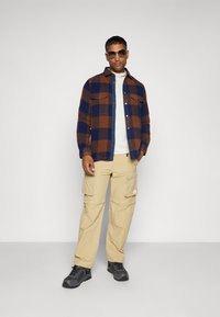 Cămașă din flanel în carouri maro și bleumarin, asociată cu o pereche de pantaloni cargo bej, pantofi gri și ochelari de soare. Design clasic și funcțional.