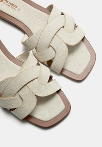 Sandales à glissière en tissu tissé beige avec un bout carré, présentant des sangles en croix et une semelle en caoutchouc brun. Matériau texturé, design plat.