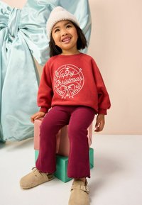 Bambino in un maglione rosso "Buon Natale", pantaloni bordeaux, scarpe beige e cappello di lana bianco, seduto su scatole regalo impilate con un grande fiocco blu.