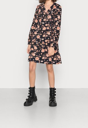 Kobieta ubrana w czarną, kwiatową sukienkę z długim rękawem, w połączeniu z czarnymi bojówkami na klamry, stojąca na jednolitym jasnym tle.