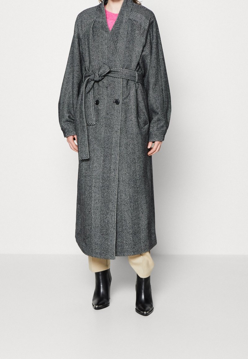 Personne portant un long manteau ceinturé noir et blanc à motif chevrons, un pantalon beige, des bottines noires et un haut rose en dessous.