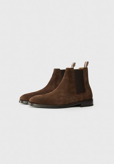 Botas Chelsea de ante marrón con paneles laterales elásticos y lengüetas. Presentan una puntera redondeada y un bajo tacón apilado, ideales para un estilo versátil.