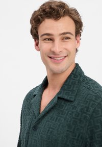 Donkergroene overhemd met kraag, voorzien van een textuurpatroon in geometrische vormen, knoopsluiting en korte mouwen. Zachte, pluizige stof.