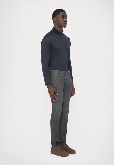 Tmavomodrá polokošeľa s dlhým rukávom a golierom, doplnená úzkymi sivými džínsami a hnedými teniskami. Jednoduchý dizajn, hladká textúra.