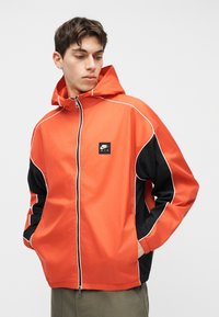 Joven con chaqueta con capucha Nike Air naranja y negra con cremallera, mirando hacia abajo, con las manos en los bolsillos, fondo neutro.