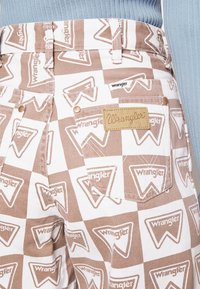 Raitapait denim ruskea ja valkoinen, jossa on kolmiomaisia "Wrangler" -logokuvioita, nahkaleima takataskussa ja metalliset niitit.