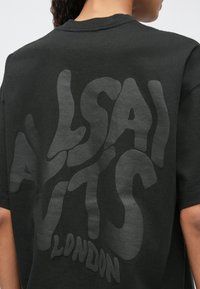 Cilvēks ar melnu T-kreklu, kuram aizmugurē ir lielas viļņotas burti, kas lasa "ALLSAINTS LONDON".