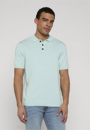 Mann trägt ein hellblaues kurzärmeliges Poloshirt mit drei schwarzen Knöpfen, kombiniert mit hellblauen Jeans, steht vor einem einfarbigen Hintergrund.