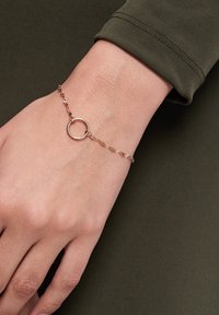 Gold-tone bracelet with a circular charm, linked chain design, and smooth texture, worn on a hand against a dark green fabric background.