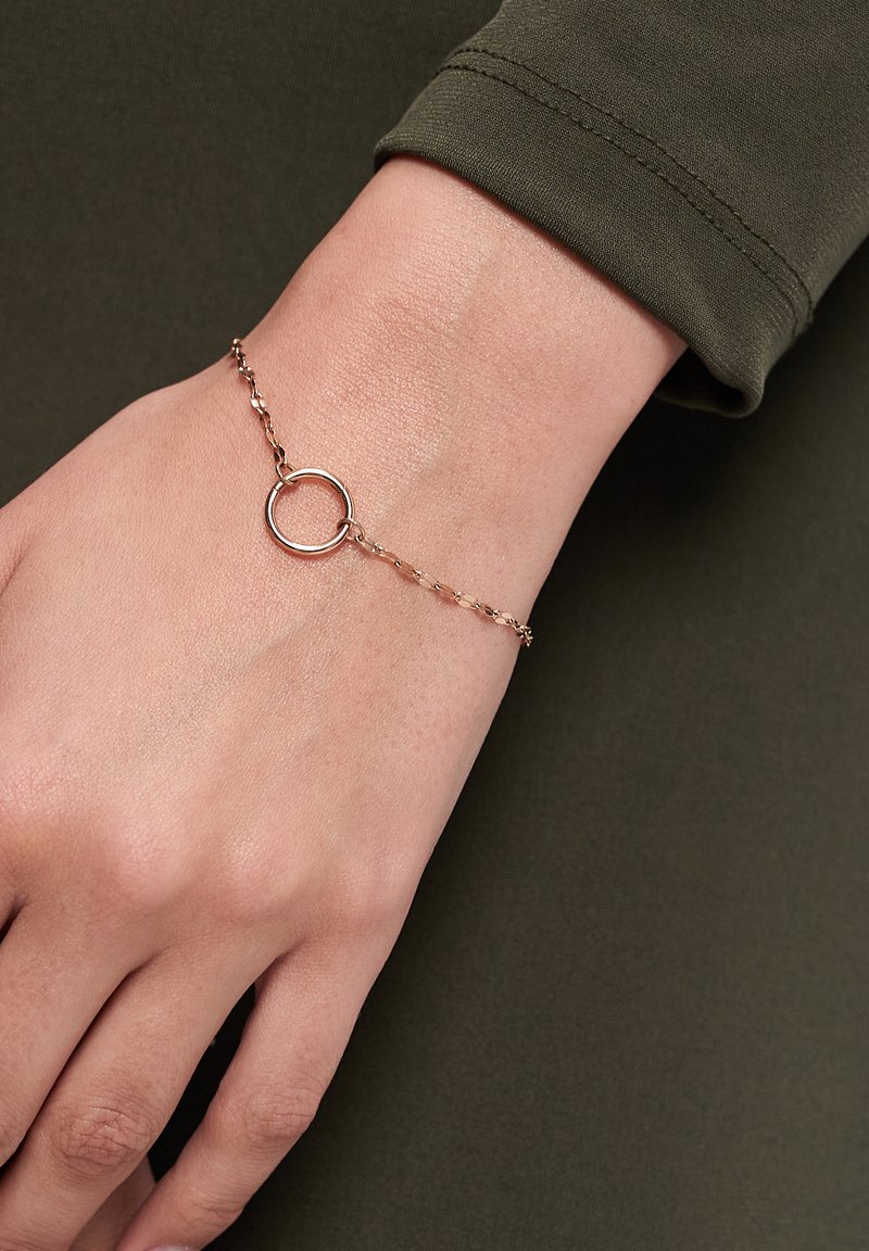 Gold-tone bracelet with a circular charm, linked chain design, and smooth texture, worn on a hand against a dark green fabric background.