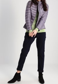 Jasnofioletowa pikowana kurtka puchowa z wysokim kołnierzem, zielony sweter, ciemne zwężane spodnie i czarne botki do kostki. Teksturowany materiał i dopasowany krój.