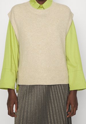 Persoon draagt een beige mouwloos vest over een limegroen shirt met lange mouwen, gecombineerd met een metalen geplooide rok, tegen een effen achtergrond.