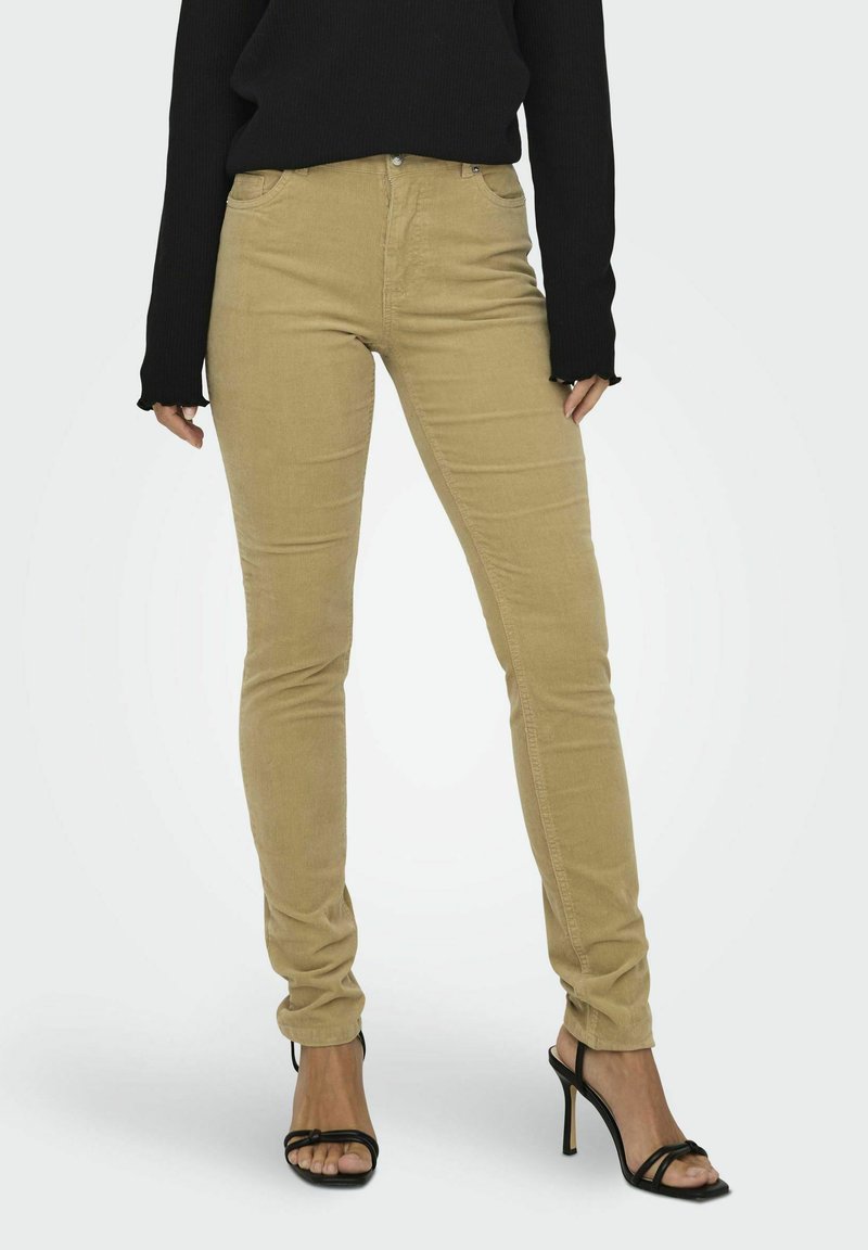 Beige, liibuvad püksid, mis on valmistatud corduroy materjalist, viie taskuga, lukuga ja kerge sädelusega, koos mustade rihmadega kontsadega.
