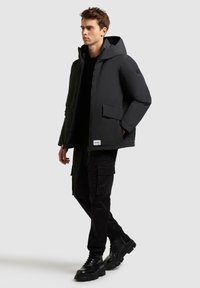 Schwarze isolierte Jacke mit Kapuze, Frontreißverschluss und zwei Seitentaschen. Über einem schwarzen Shirt getragen und kombiniert mit schwarzen Cargo-Hosen und Stiefeln.