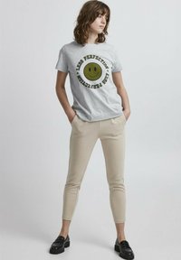 Grijze t-shirt met een grote gele glimlach en zwarte tekst met "Less Perfection" in een cirkelvormig patroon. Beige slim-fit broek en zwarte instaplaarzen.
