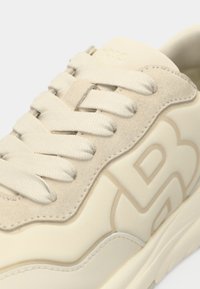 Baskets beiges avec une empeigne en suède texturé, des lacets plats et un logo en relief discret sur le côté. Silhouette arrondie et semelle lisse.