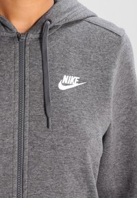 Grå zip-up hoodie i mjukt tyg, med en vit logotyp på vänster bröst och dragsnören på huvan.