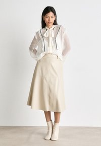 Cremefarbene Bluse aus strukturiertem Stoff mit schwarzem Besatz, kombiniert mit einem beigen A-Linien-Rock und cremefarbenen Ankle Boots, steht vor einem neutralen Hintergrund.