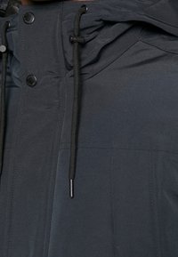 Donkergrijze jas met zwarte trekkoorden, zwarte drukknopen en zichtbare stiksels op de stofpanelen en kap.