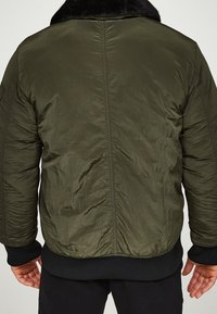 Chaqueta de plumas verde oliva con cuello de piel sintética negra, que presenta un exterior texturizado, puños acanalados y costuras en la parte posterior para mayor estructura.