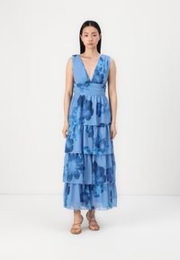 Zila ziedu maksi kleita ar dziļu V veida kaklu, kārtotiem volāniem un pieguļošu vidukli. Audums izskatās viegls un plūstošs.