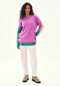 Žena s dlhými kučeravými vlasmi, oblečená v ružovo-zelenom svetri s farebnými blokmi, bielych nohaviciach a hnedých turistických topánkach, stojaca s jednou rukou na boku.