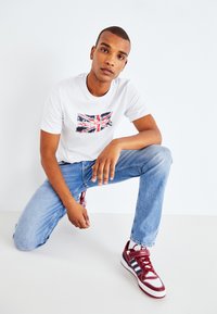 Witte t-shirt met een Union Jack graphic, gecombineerd met lichtblauwe jeans en bordeauxrode sneakers met witte en marineblauwe accenten.
