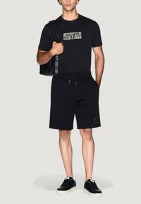 Camiseta negra con un logotipo texturizado, combinada con pantalones cortos negros con una cintura elástica y un acento de logotipo. Llevada con zapatillas negras.