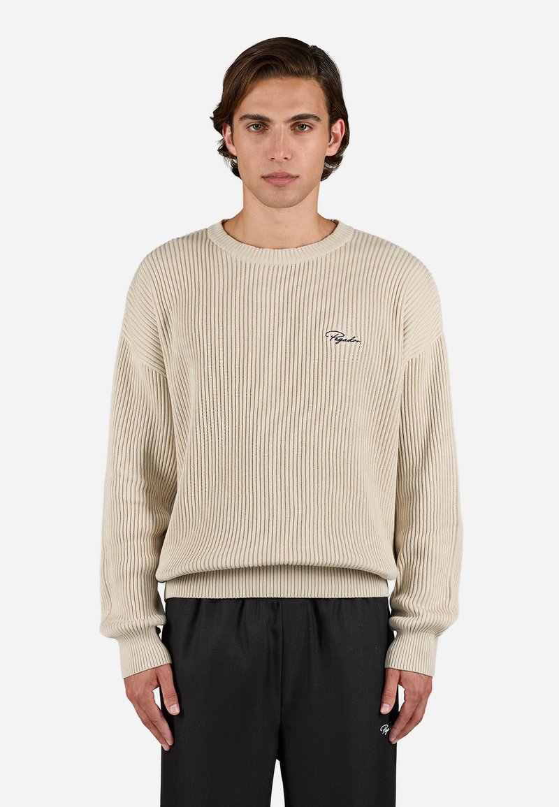 Uomo con un maglione a coste beige e pantaloni neri, in piedi con le braccia rilassate, rivolto in avanti su uno sfondo chiaro e semplice.