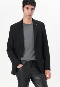 Čierny blazer s jedným gombíkom, klopovými reverzami a náprsníkom, nosený cez šedý sveter; spárovaný s čiernymi koženými nohavicami.