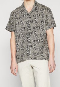 Košile s krátkým rukávem v světle béžové a černé barvě se vzorem obdélníkových a oválných tvarů, se rozšířeným límcem a zapínáním na knoflíky.