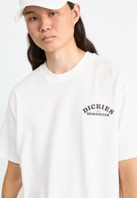 Hvit bomull T-skjorte med rund hals, med et trykt "Dickies Workwear" logo i svart og rosa. Korte ermer, avslappet passform.