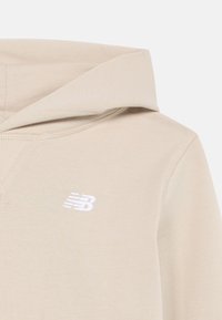 Beige színű kapucnis pulóver, fehér New Balance logóval hímzett a mellkasán, részletes varrással és szövet textúrával.