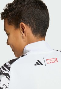 Nuorten urheilutakki, jossa valkoinen runko, mustat yksityiskohdat, logot sekä korkea kaulus. Hihoissa on näyttävä graafinen kuvio.