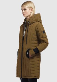 Donna che indossa un lungo cappotto trapuntato verde oliva con cappuccio, polsini neri, tasche laterali e zip con logo, voltata verso sinistra su uno sfondo semplice.