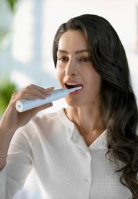 Brosse à dents électrique avec un corps blanc allongé et une tête de brosse fine tenue dans une main, utilisateur brossant ses dents. L'arrière-plan présente une lumière douce.