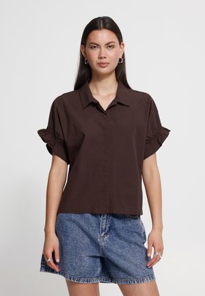 Femme aux longs cheveux foncés porte un chemisier à manches courtes marron foncé avec des poignets volantés et un short en jean bleu sur un fond clair uni.