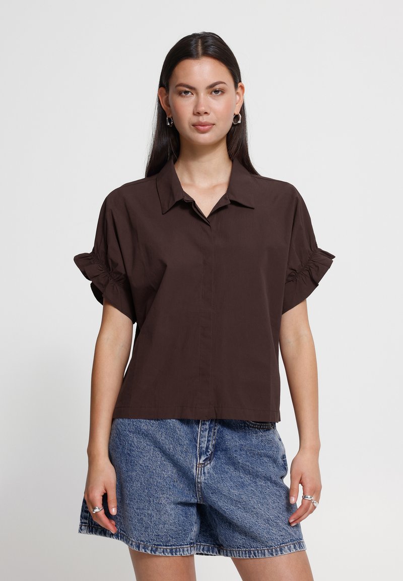 Femme aux longs cheveux foncés porte un chemisier à manches courtes marron foncé avec des poignets volantés et un short en jean bleu sur un fond clair uni.