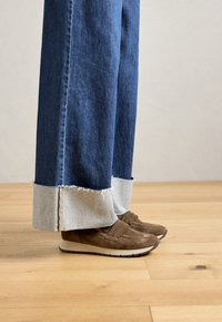 Dark blue wide-leg jeans with frayed cuffs rolled up, worn with brown suede slip-on shoes on a wooden floor.