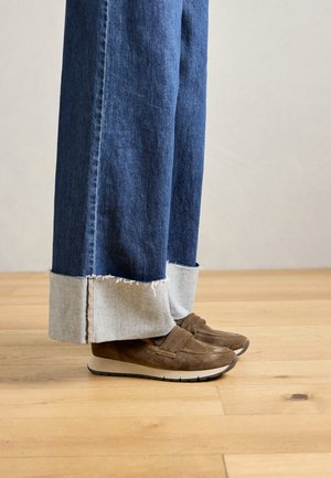 Tumedad sinised laiad püksid, mille alumised servad on kärbitud ja üles keeratud, kantud pruunide sametiste libisemisjalanõudega puidust põrandal.