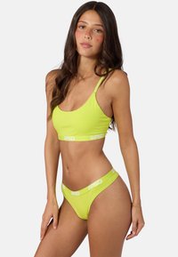 Femme aux longs cheveux bruns portant un soutien-gorge de sport vert citron et un sous-vêtement assorti avec des logos PSD blancs sur les bandes élastiques.